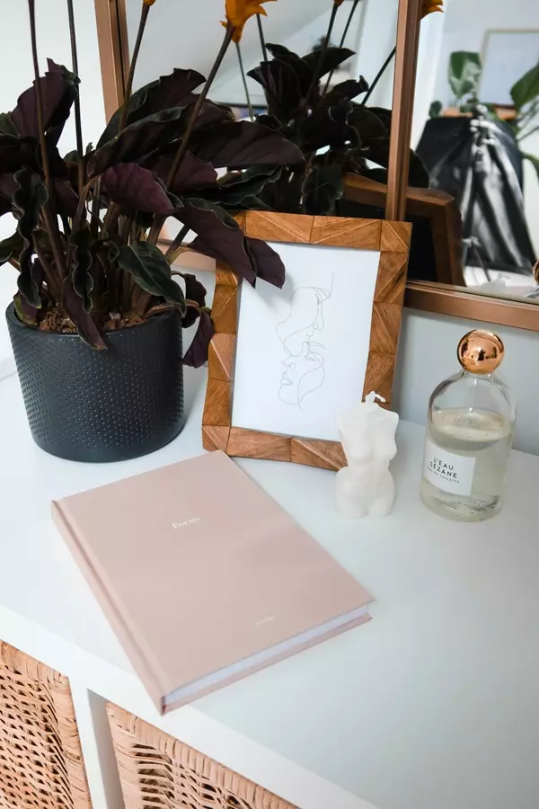 Tavolo bianco con pianta, cornice senza foto, diario colore rosino e botteggino di profumo con a fianco una candela bianca
